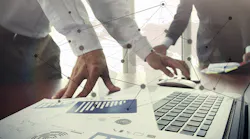 Business documents on office table with smart phone and digital tablet and stylus, and two colleagues discussing data in the background.